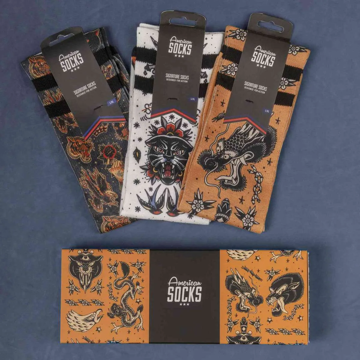Drei verpackte American Socks mit Tattoo-Motiven und eine passende Geschenkbox liegen auf einem blauen Untergrund.