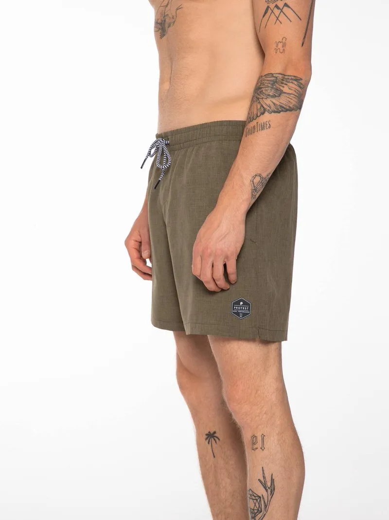 Ein Mann in grünen Badeshorts mit Kordelzug und "Protest"-Logo, Seitenansicht, Tätowierungen an Armen und Beinen.