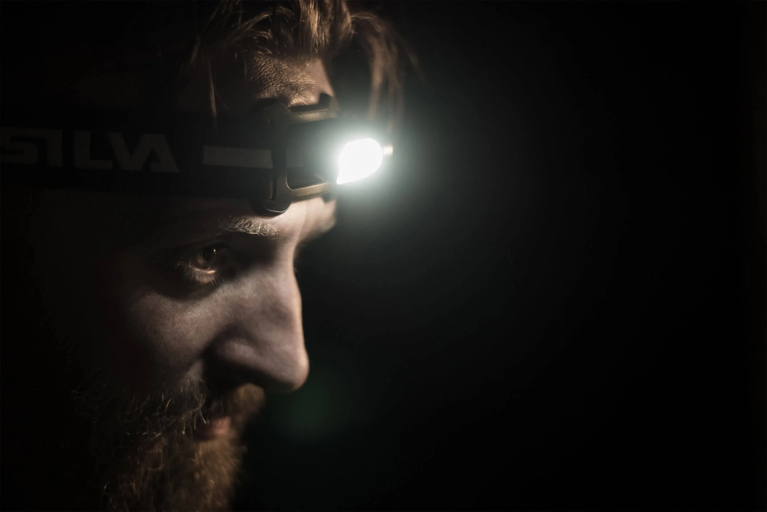 Profil eines Mannes mit Bart, der eine eingeschaltete Silva Scout 3 Stirnlampe trägt.
