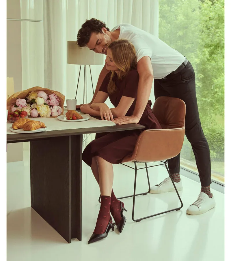 Paar am Tisch; Frau mit Velour Beat Socken Lipstick und Samtherzen, Mann umarmt sie.