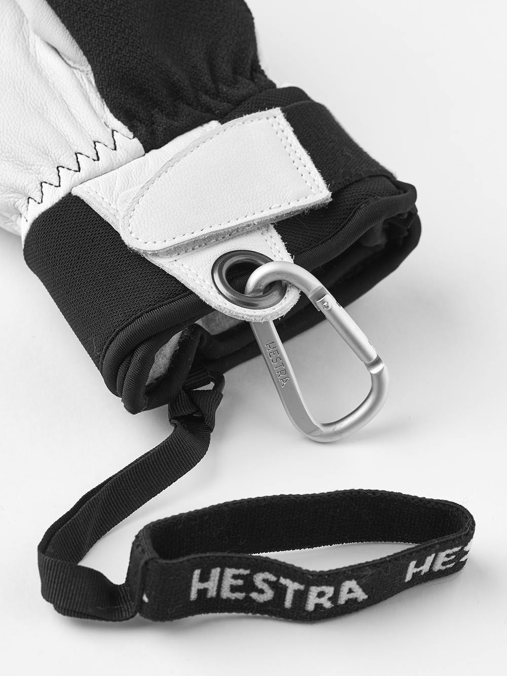 Nahaufnahme eines Handschuhs in Schwarz und Weiß mit Karabiner und einem Band mit dem Wort "HESTRA".