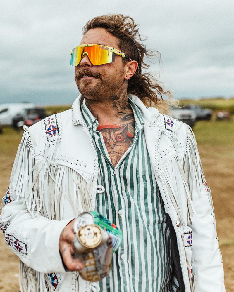Mann mit bunten 1993 2000's Sonnenbrille, Fransenjacke hält Getränke. Tattoos auf Hals und Brust.