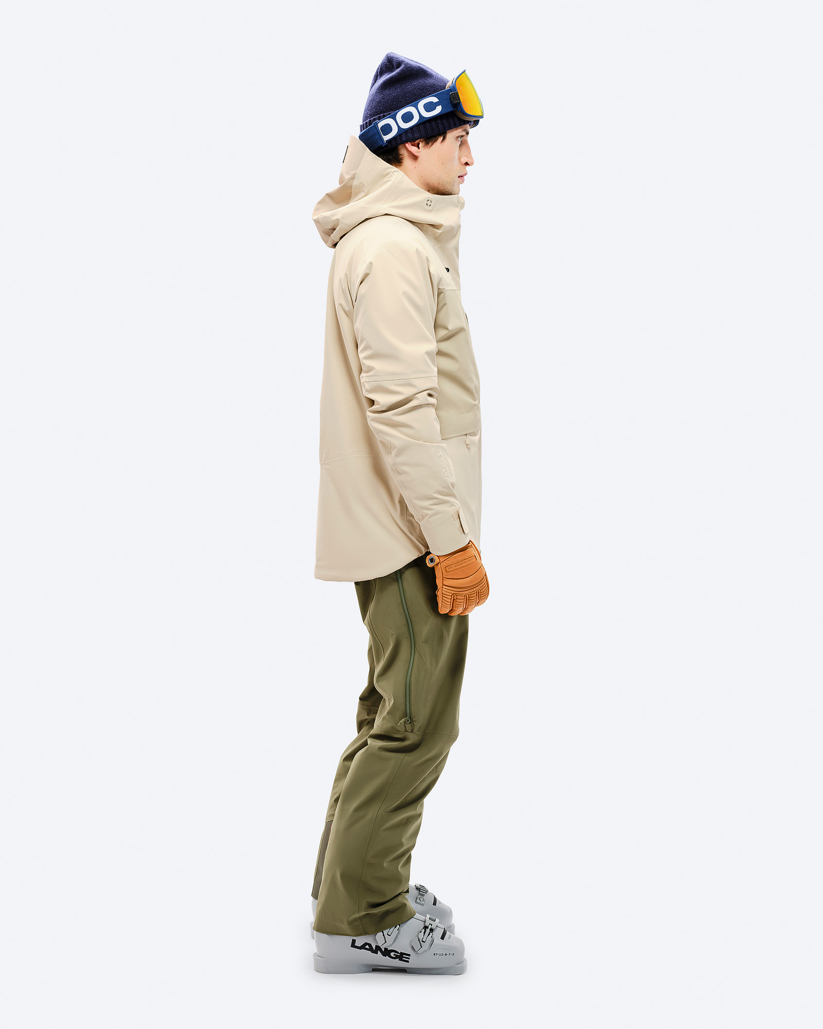 Ein Mann in Ski-Ausrüstung von der Seite: Beige Jacke, grüne Hose, Skischuhe, Mütze und Brille.