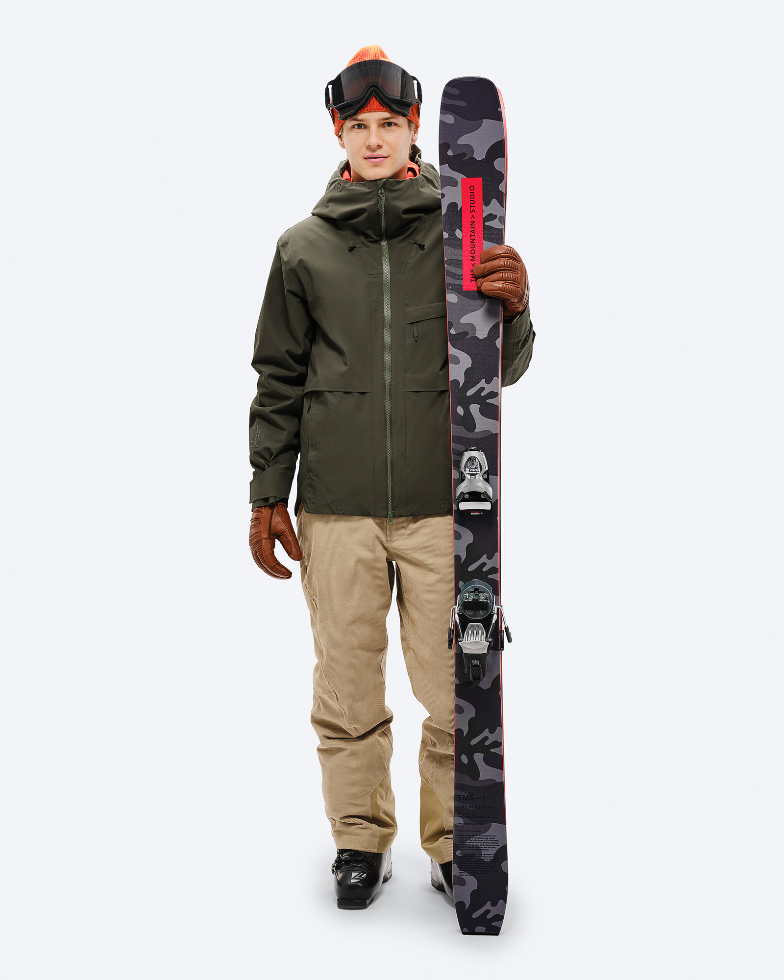 Model mit S-3 Carv Insulated Stretch Jacket in Grün hält Ski mit Tarnmuster, trägt Mütze und Skibrille.