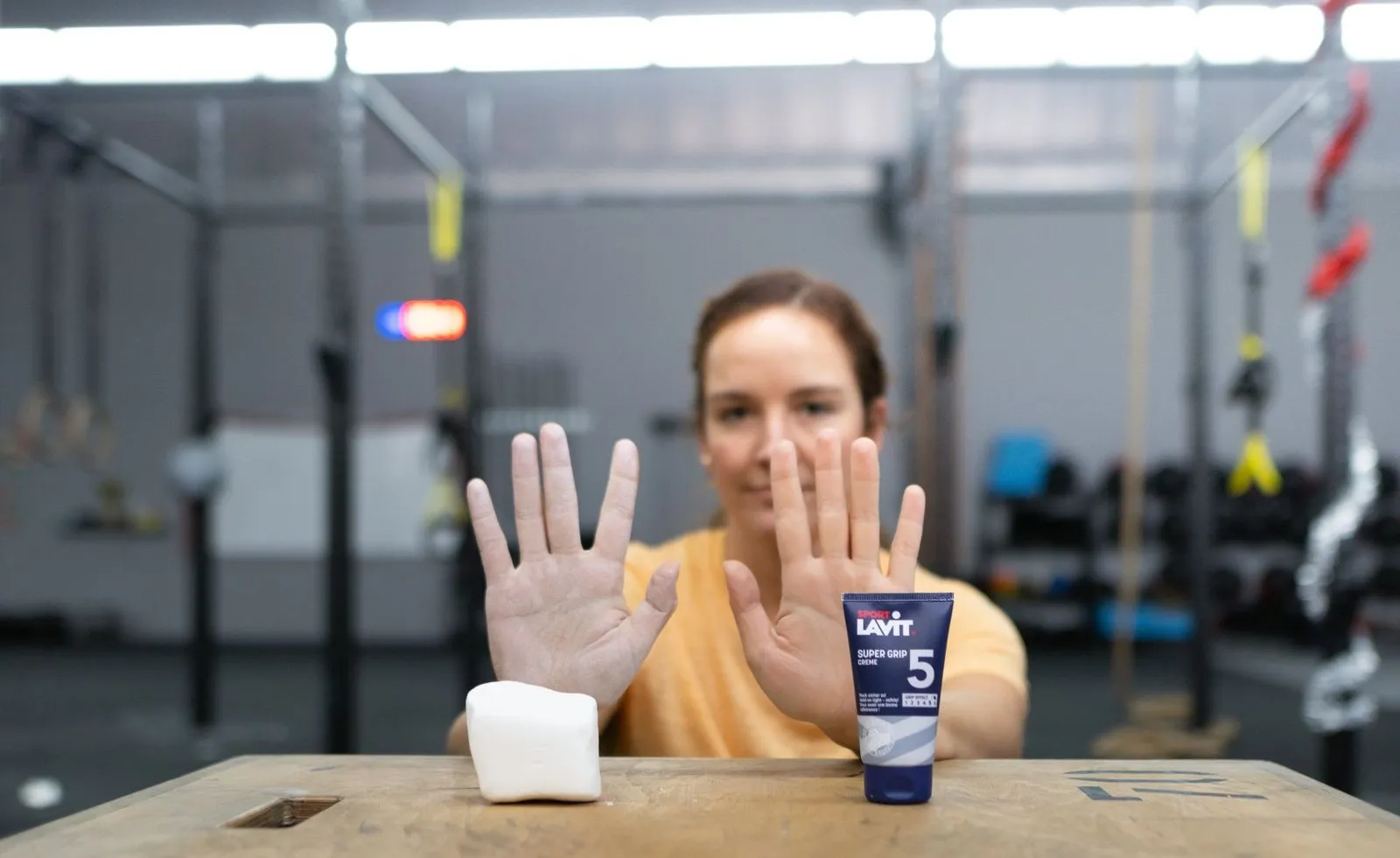 Frau zeigt Hände, Tube SPORT LAVIT Super Grip Creme und ein Magnesium Block auf Holztisch im Fitnessstudio.
