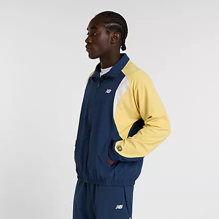 Ein Mann mit marineblauer New Balance In Game Track Jacket mit gelben Ärmeln, Reißverschluss und Seitentaschen.