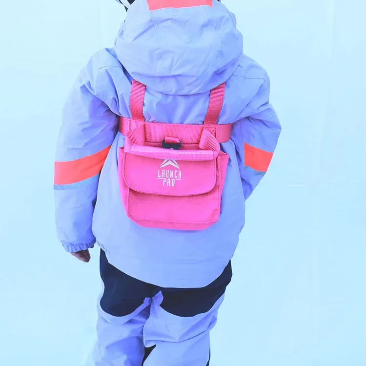 Von hinten: Kleinkind in lavendelfarbener Skijacke und -hose mit leuchtend orangefarbenen Streifen an den Ärmeln. Am Rücken ein pinkfarbenes Ski-Gurtzeug mit Beutel und weißem Logo „LAUNCH PAD“. Gurte über Schultern und Taille.