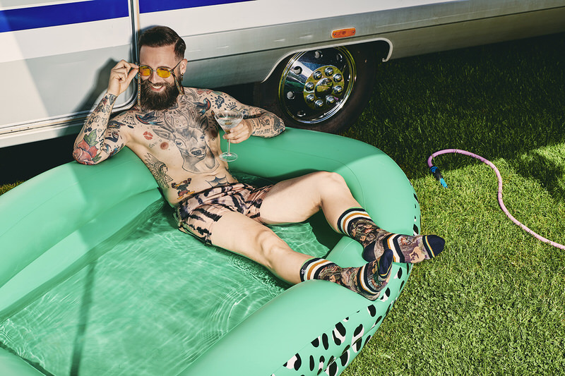 Entspannt im Pool Mann mit Tattoos, Badehose und Hawaii Blues Socken entspannt im Pool mit Drink vor einem Wohnwagen.