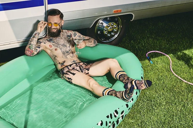 Mann mit Tattoos, Badehose und Hawaii Blues Socken entspannt im Pool mit Drink vor einem Wohnwagen.