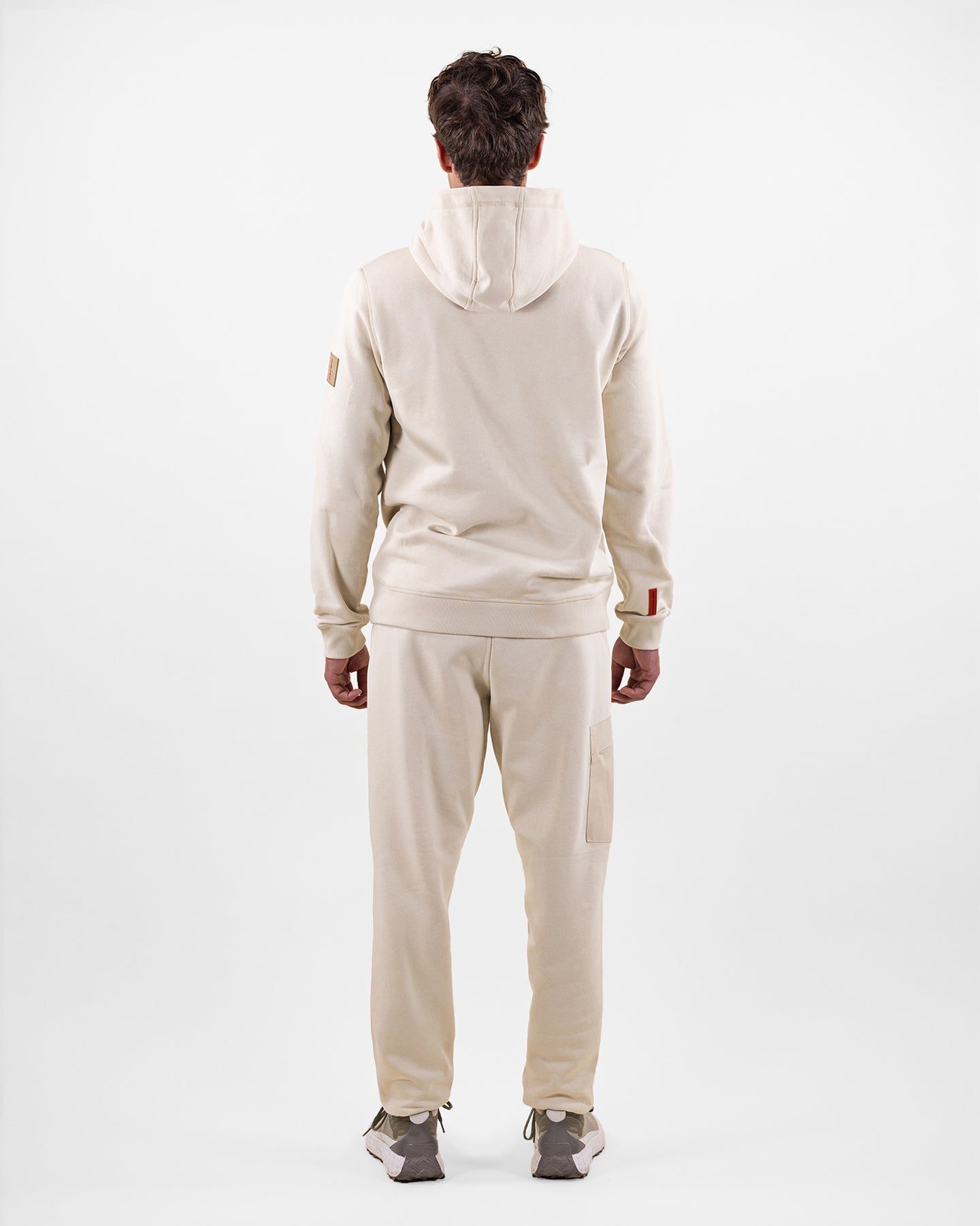 Rückansicht eines Mannes in beigem Hoodie-Anzug und Turnschuhen, vor weißem Hintergrund.