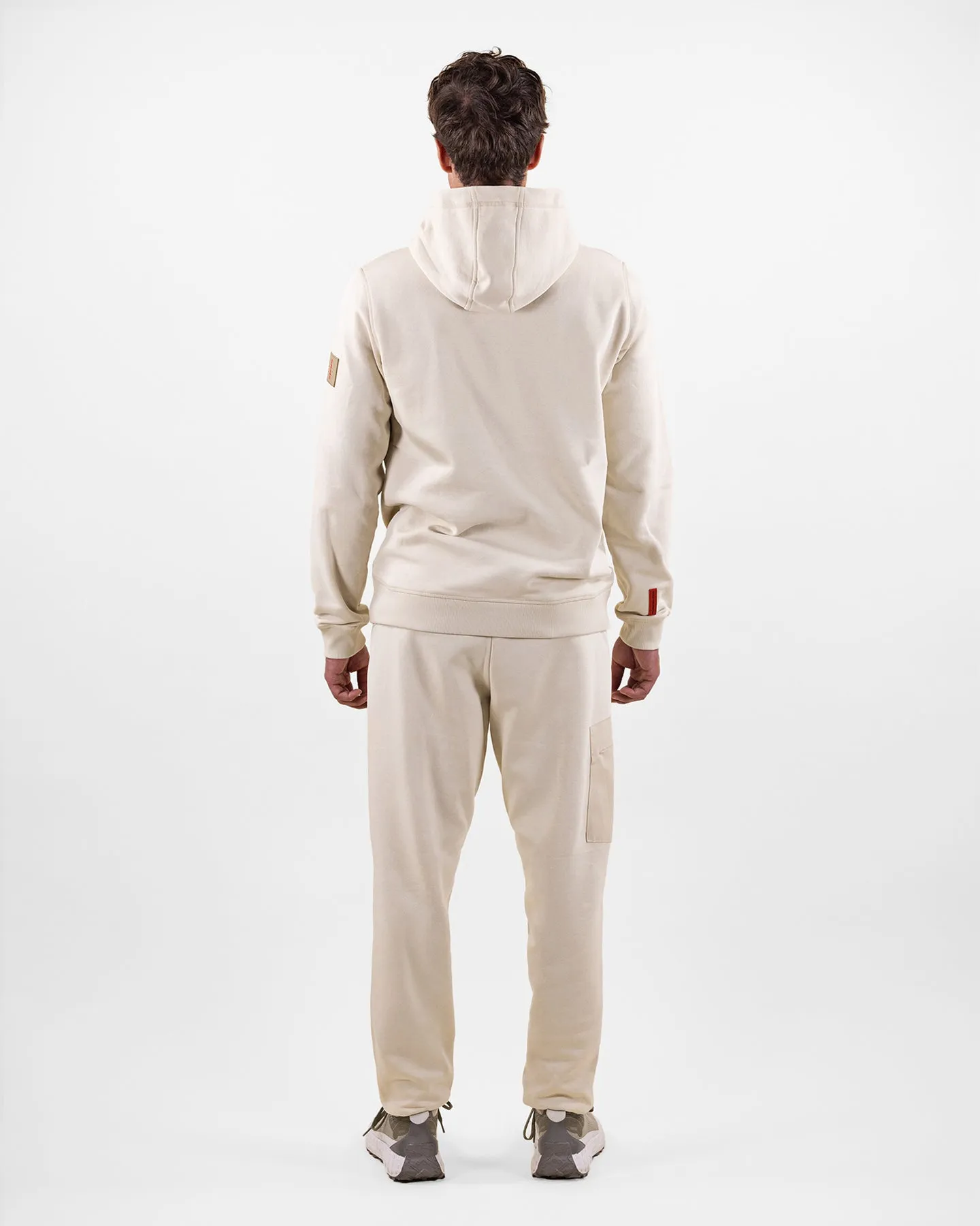 Rückansicht eines Mannes in beigem Hoodie-Anzug und Turnschuhen, vor weißem Hintergrund.