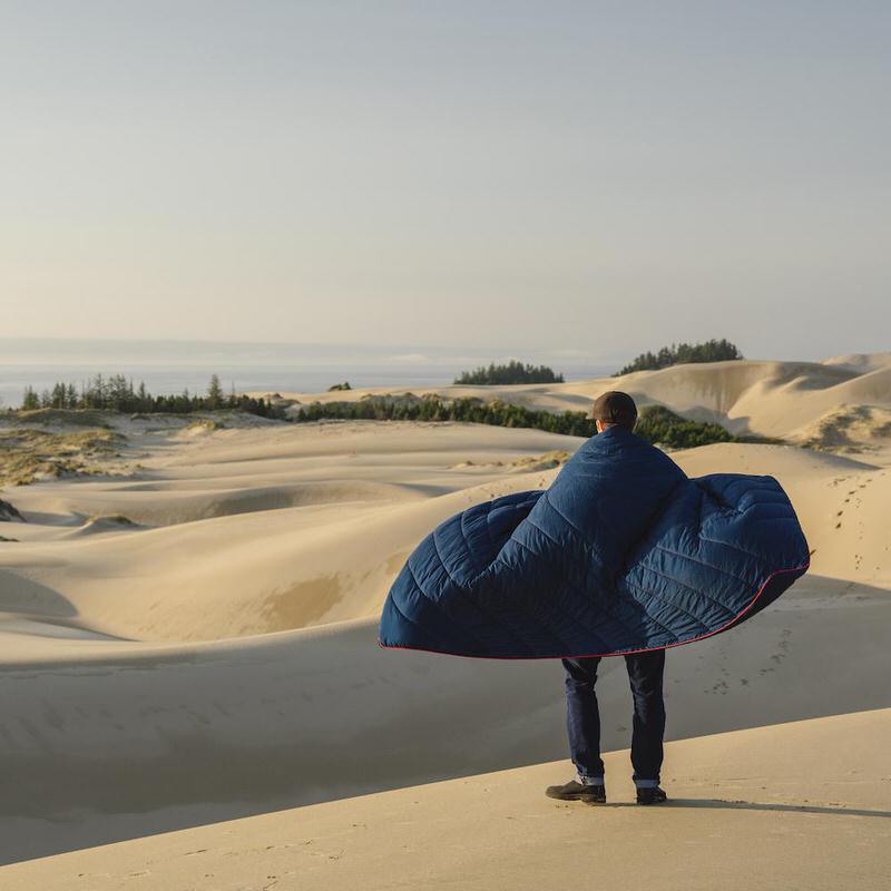 Mann steht mit Deepwater Rumpl Decke in Dünenlandschaft, Blick aufs Meer. Die Puffy Blanket ist wie ein Umhang umgelegt.