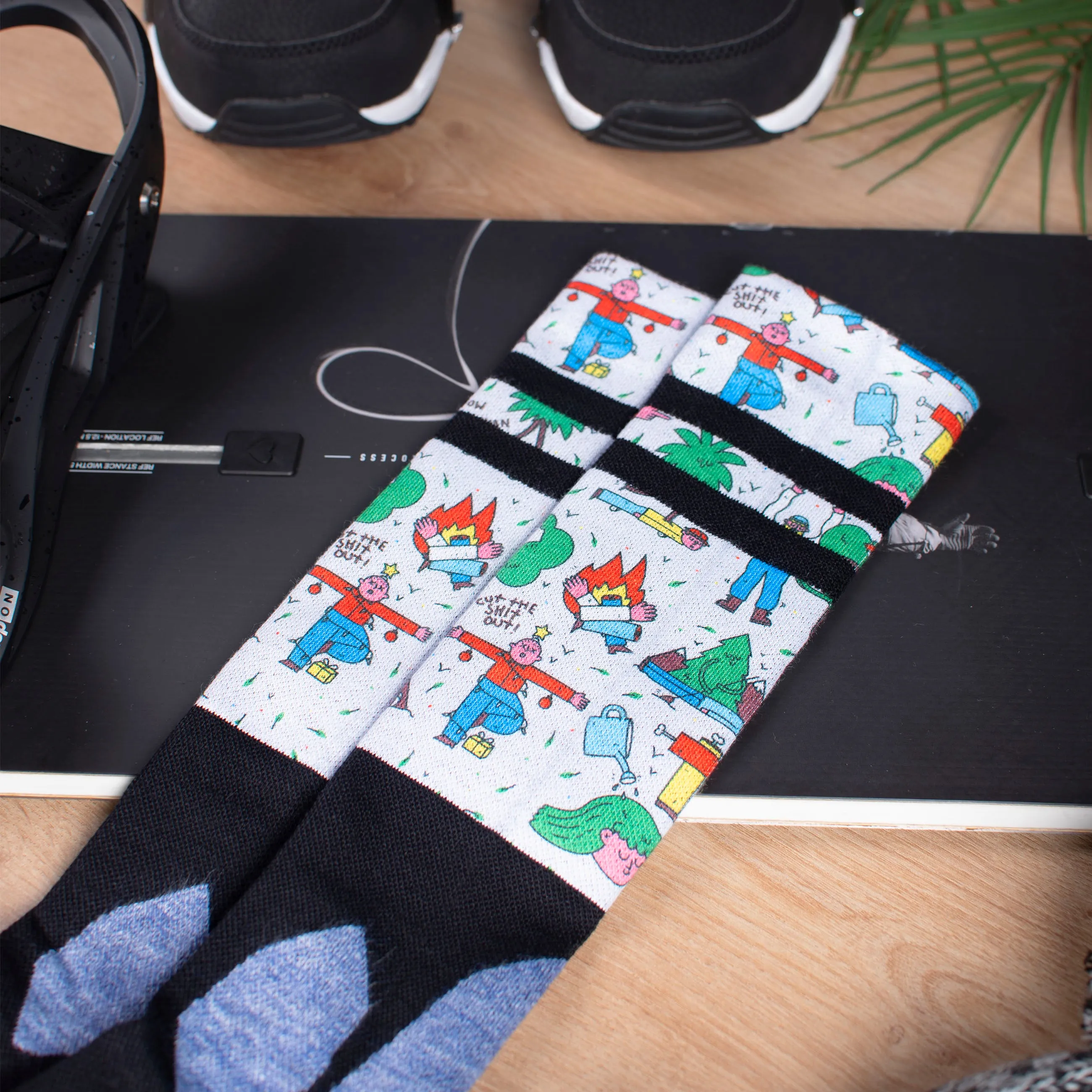 Kniehohe Snow Socks auf Snowboard, Motiv mit Comicfiguren, schwarz-weiß gestreift, dazu Snowboard Boots und ein Blatt.