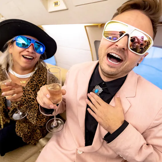 Zwei Personen mit Flight Optics Sonnenbrillen, Champagnergläsern, feiern. Der Mann trägt ein rosa Jackett.