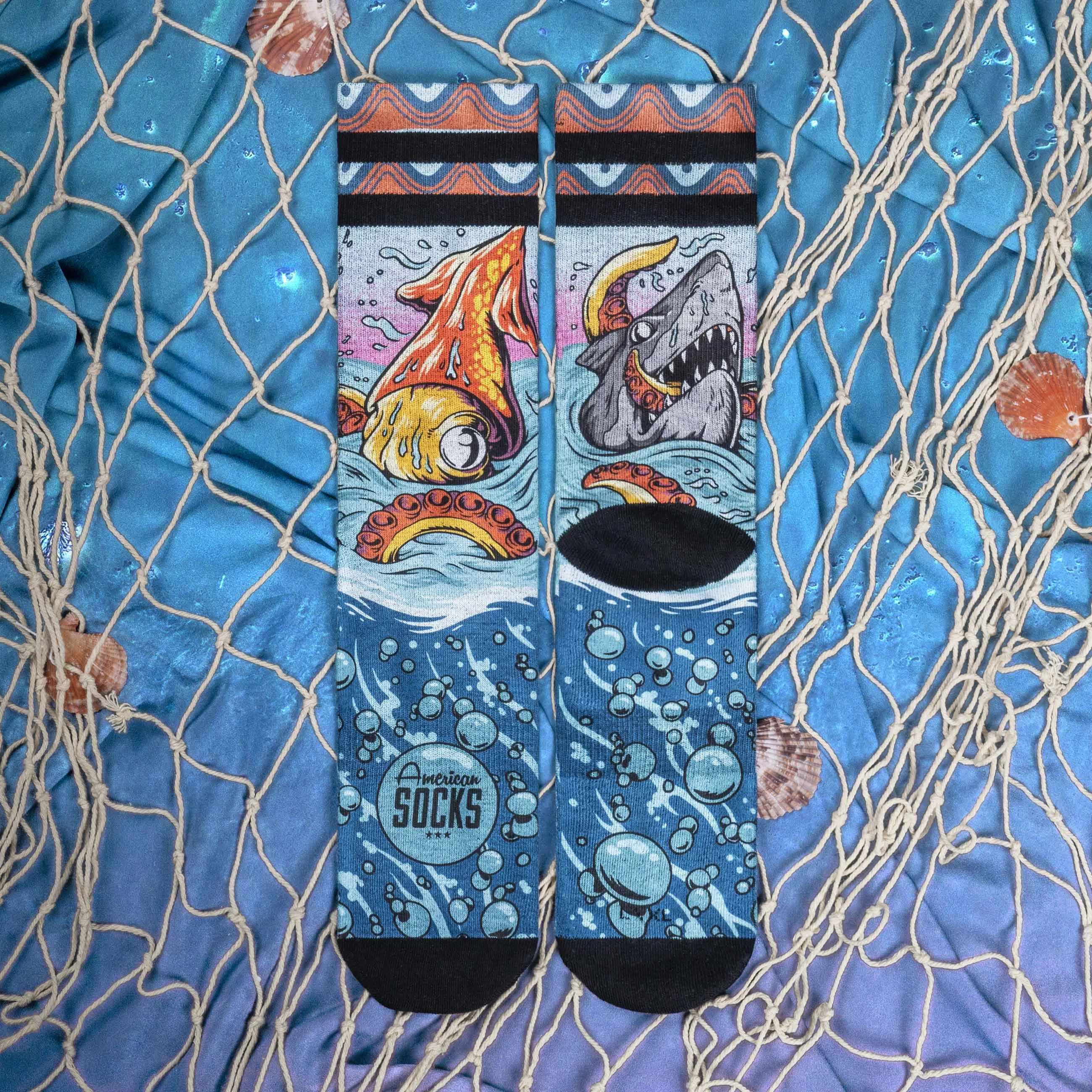 Zwei 'Seamonsters Mid High' Socken mit Tintenfisch, Hai, Blasen und 'American Socks' Logo auf blauem Stoff mit Muscheln und Fischernetz.