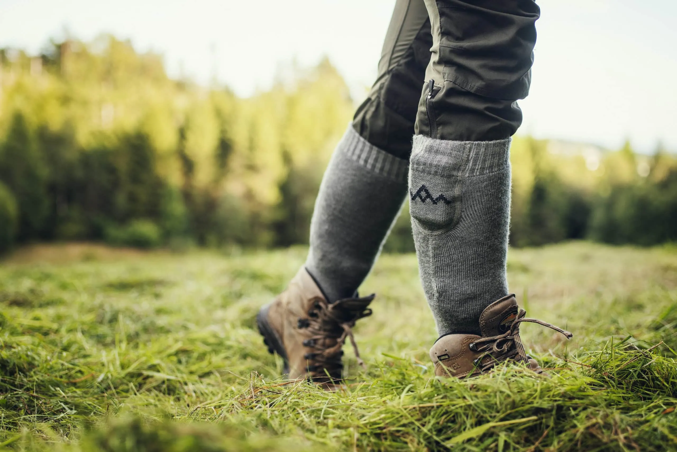 Person in Wanderschuhen und Everyday Heizsocken V2 auf einer grünen Wiese. Nahaufnahme.