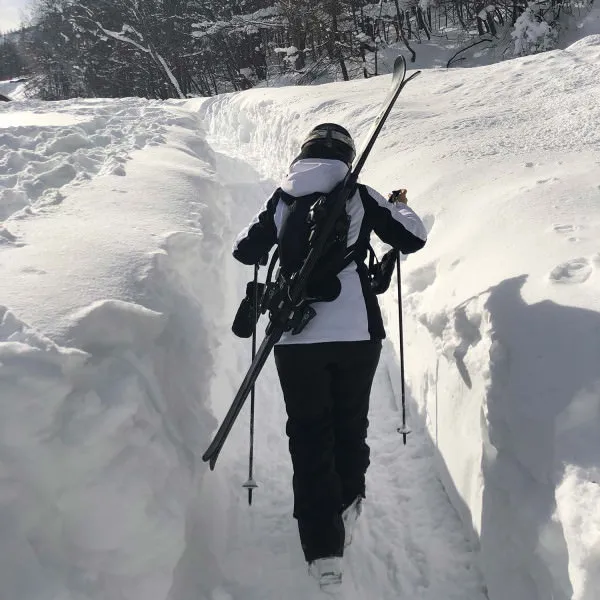 Person mit Skiern auf dem Rücken und Skistock in der Hand geht durch den Schnee. Skiback ist sichtbar.