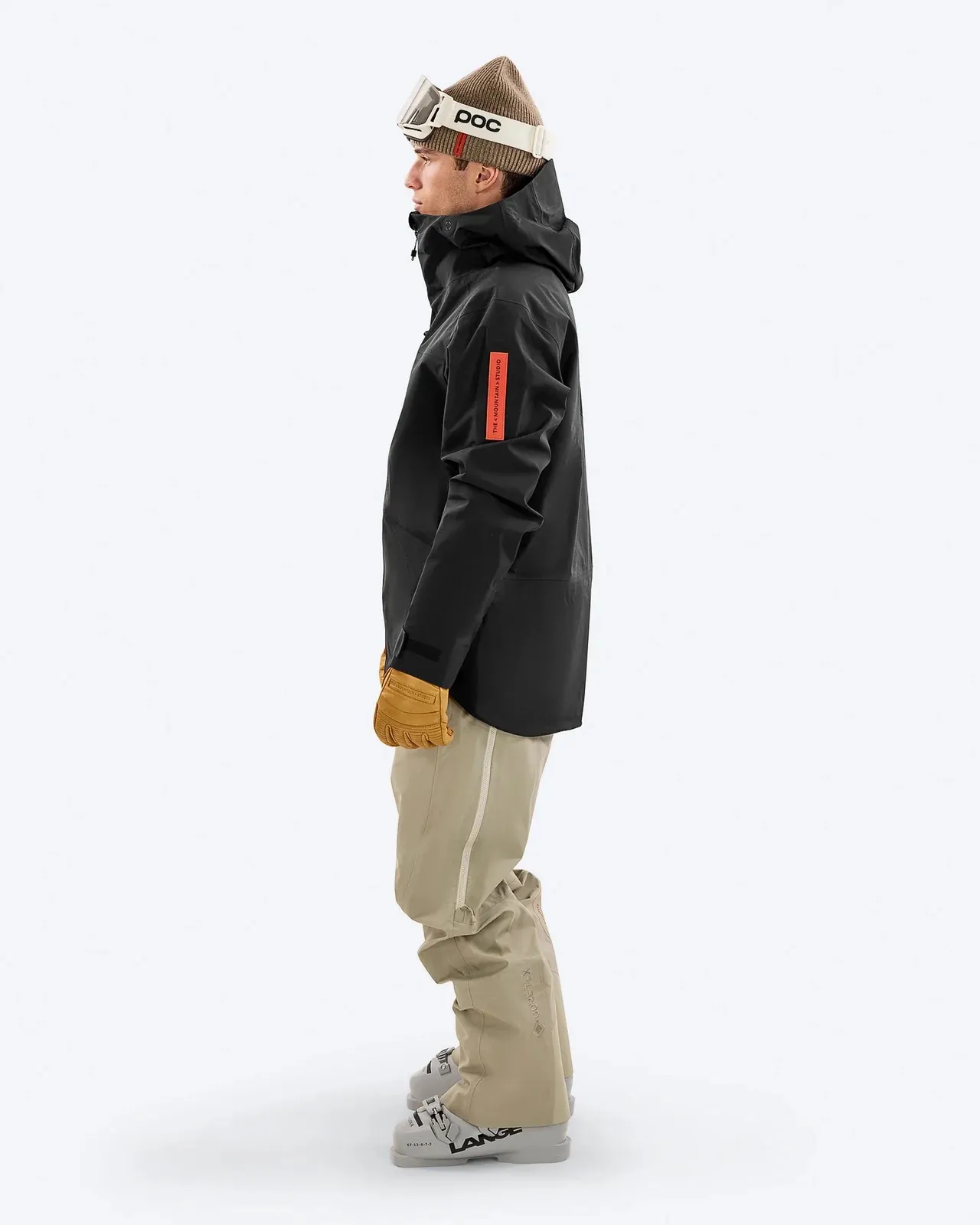 Person in Skikleidung, seitlich. Sie trägt eine schwarze Jacke, beigefarbene Hose, Mütze und Skibrille. Skischuhe sind sichtbar.
