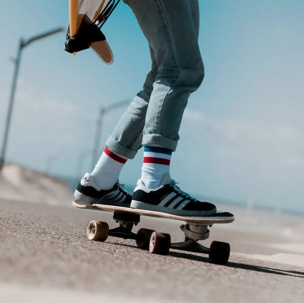 Person auf Skateboard mit American Pride Mid High Socken, Jeans und Turnschuhen vor blauem Himmel.