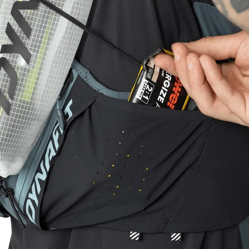 Nahaufnahme eines schwarzen Dynafit-Laufrucksacks. Eine Hand nimmt einen Powerbar-Energieriegel aus einer Seitentasche. Die perforierte Tasche zeigt kleine gelbe Punkte. Eine Trinkblase mit weißem Netzmuster ist sichtbar.