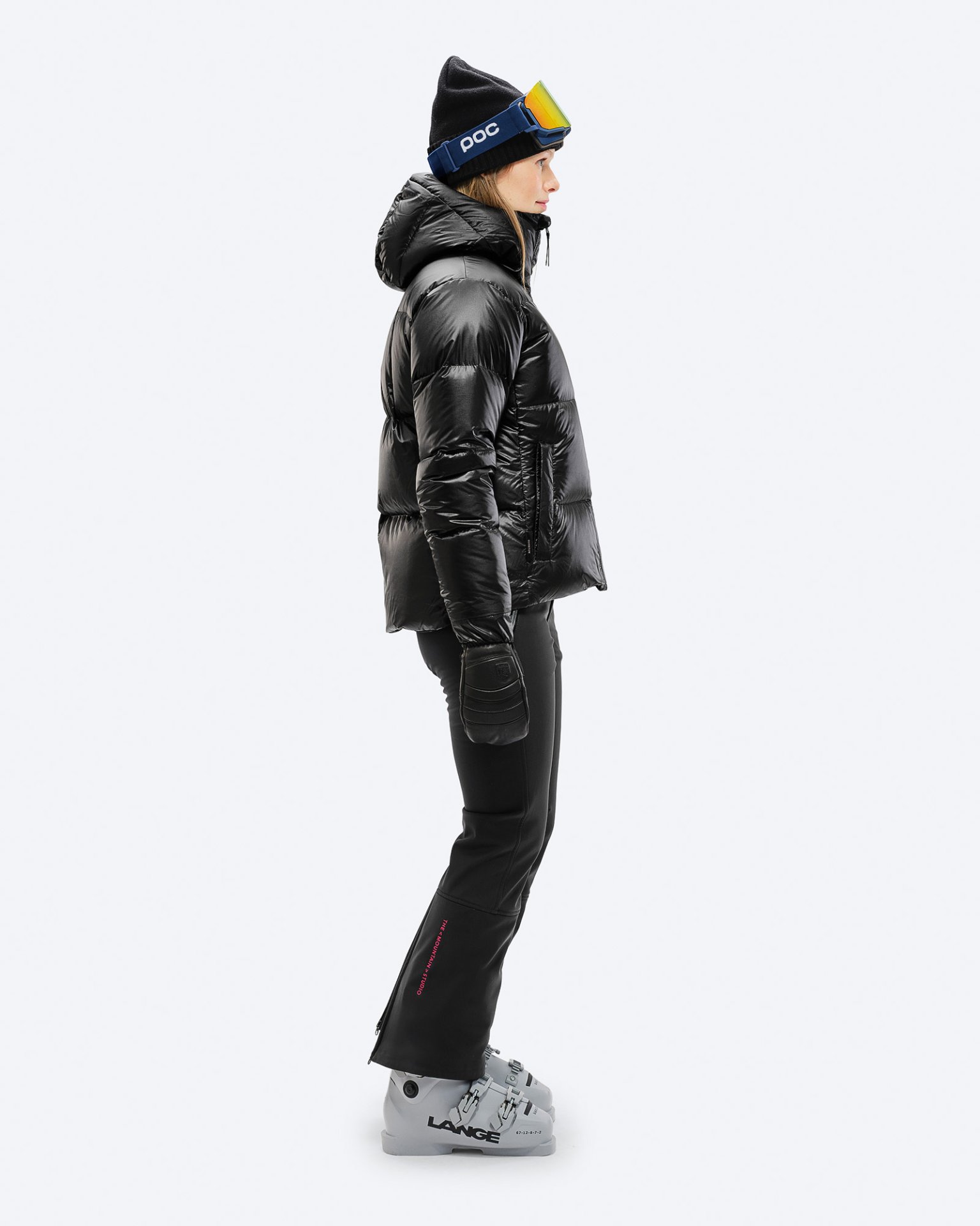 Frau in schwarzer Skihose und Jacke von der Seite, mit Skischuhen, Mütze und Skibrille.