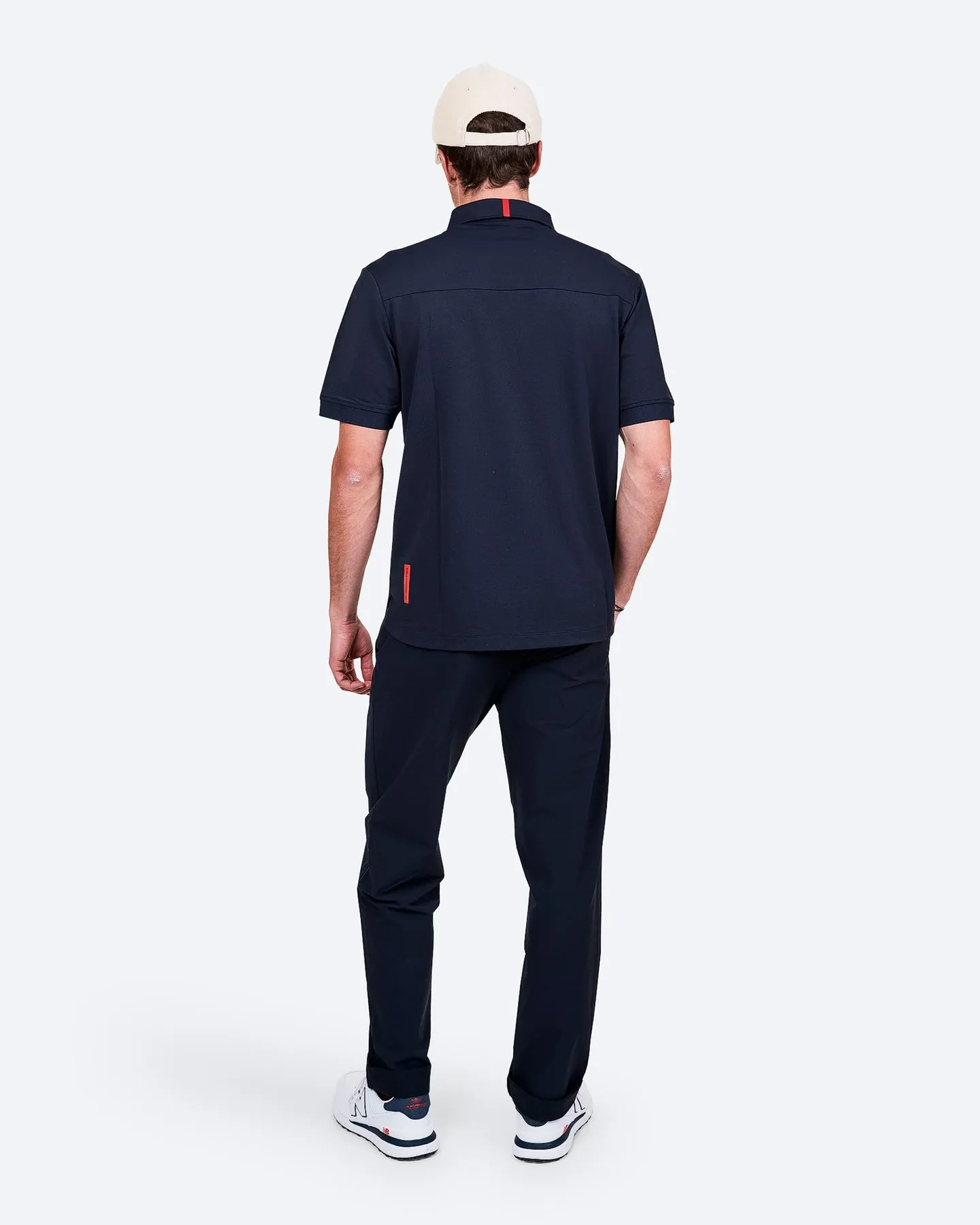 Rückansicht eines Mannes in marineblauer Kleidung, beiger Kappe und weißen Turnschuhen auf weißem Grund.