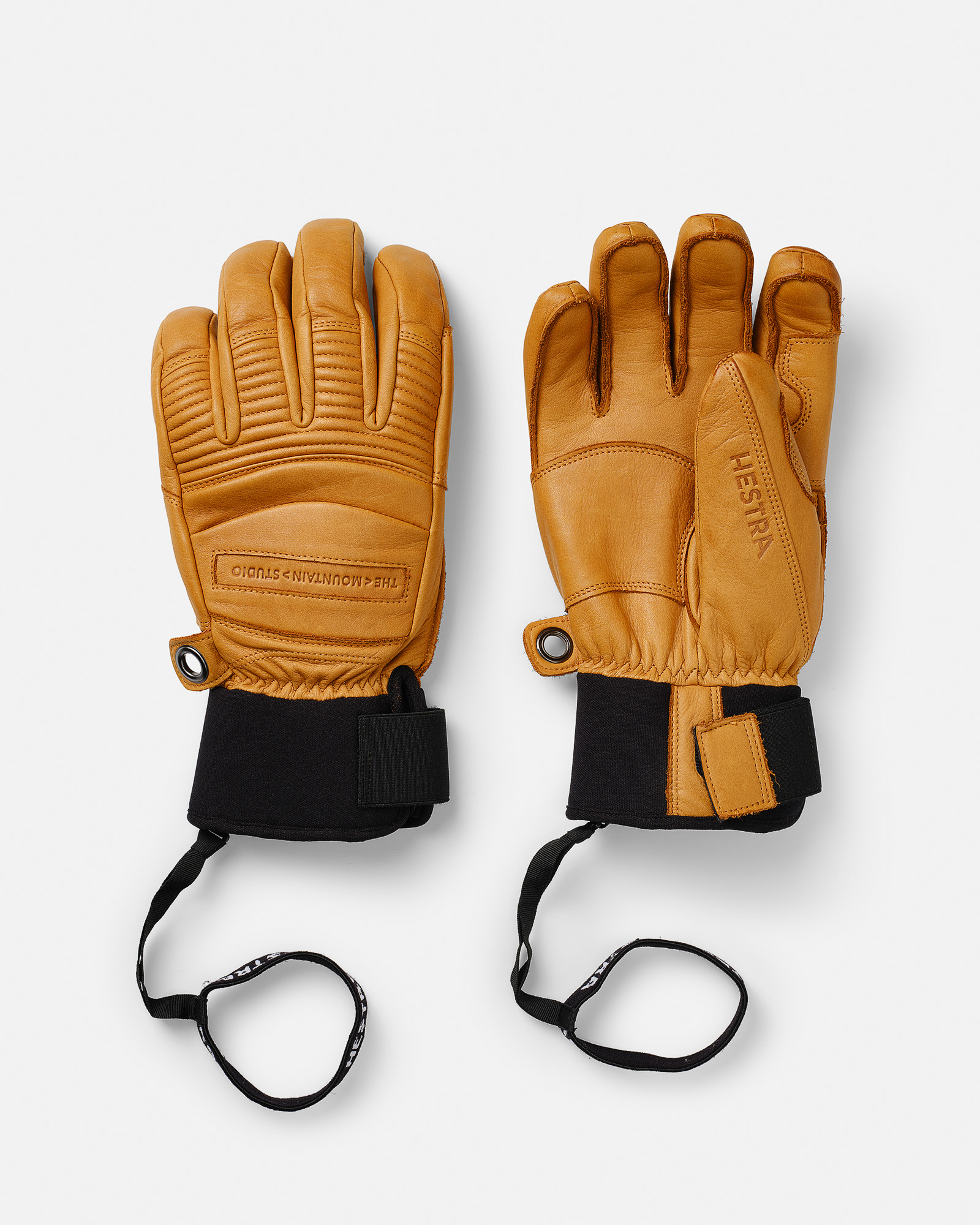 Zwei HESTRA® Lederhandschuhe in Sandfarbe mit schwarzen Neoprenmanschetten und Sicherheitsriemen auf weißem Hintergrund.