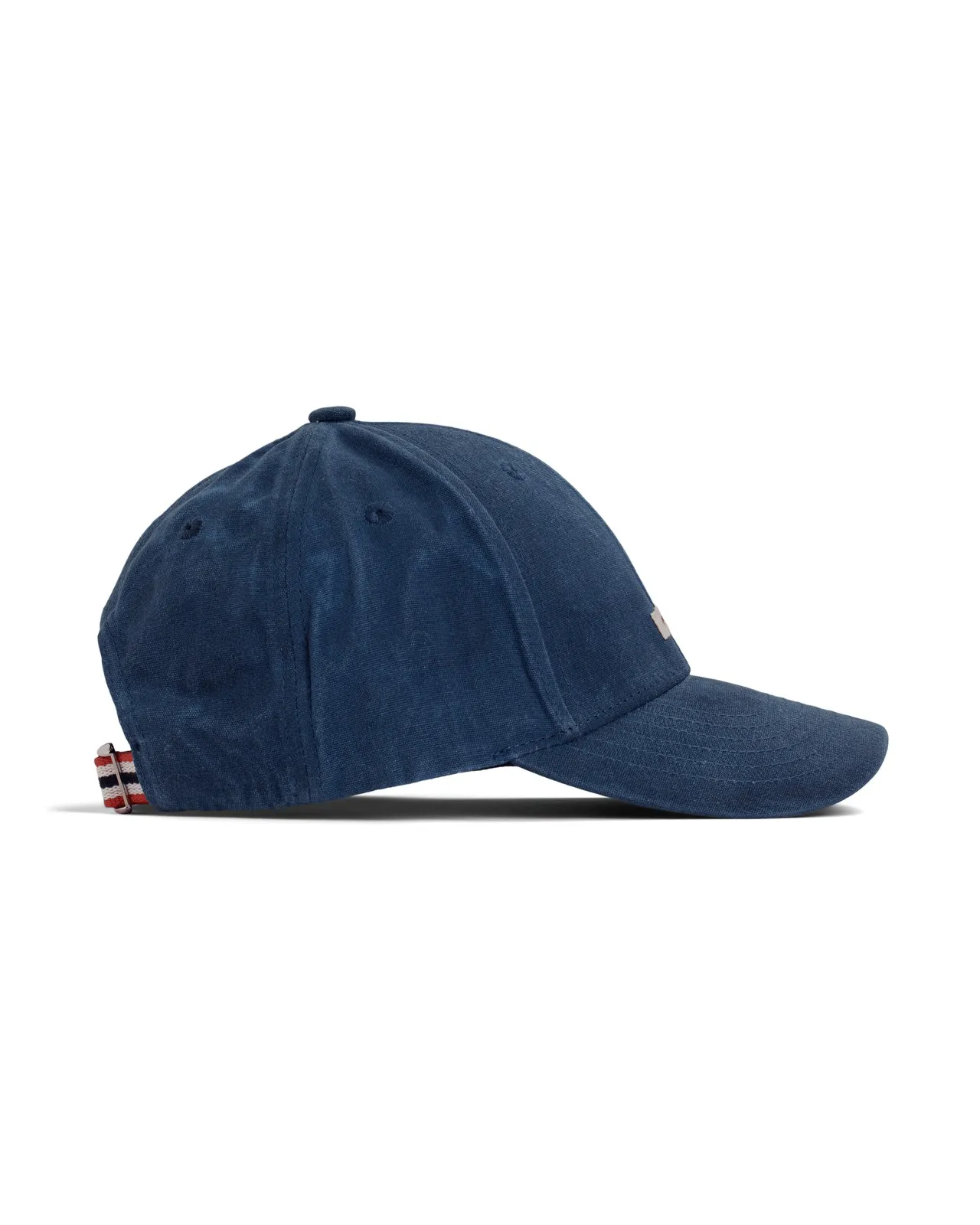 Seitliche Ansicht einer marineblauen Amundsen Waxed Cotton Cap vor einem weißen Hintergrund. Die Kappe hat einen Knopf oben, einen gebogenen Schirm und einen Riemen mit rot-weiß-blauen Streifen am Hinterkopf zur Größenverstellung.