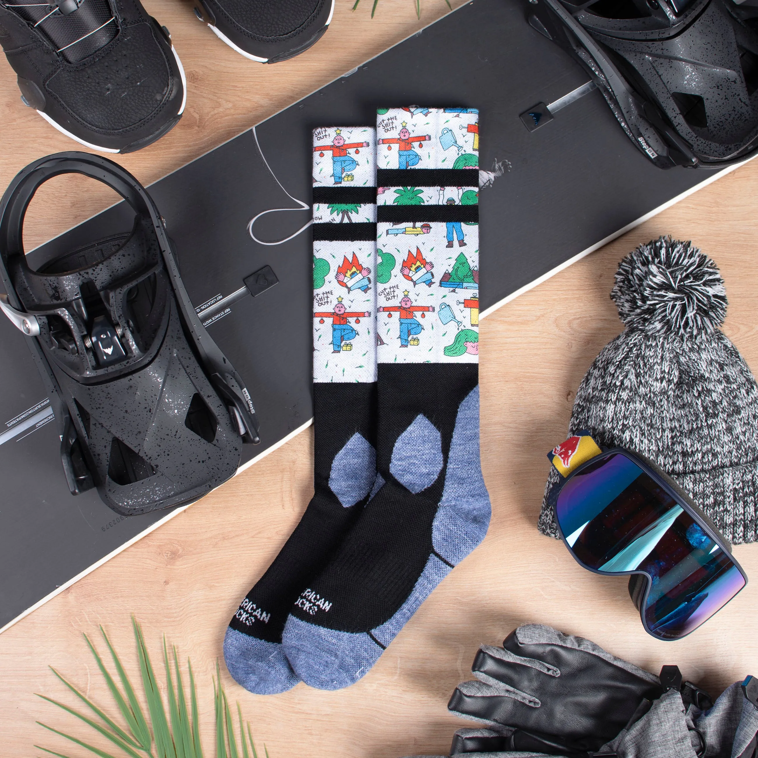Snow Socks "Leaf Me Alone" auf Snowboard mit Boots, Mütze, Handschuhe und Skibrille auf Holzuntergrund. Socken mit Comic Motiven.