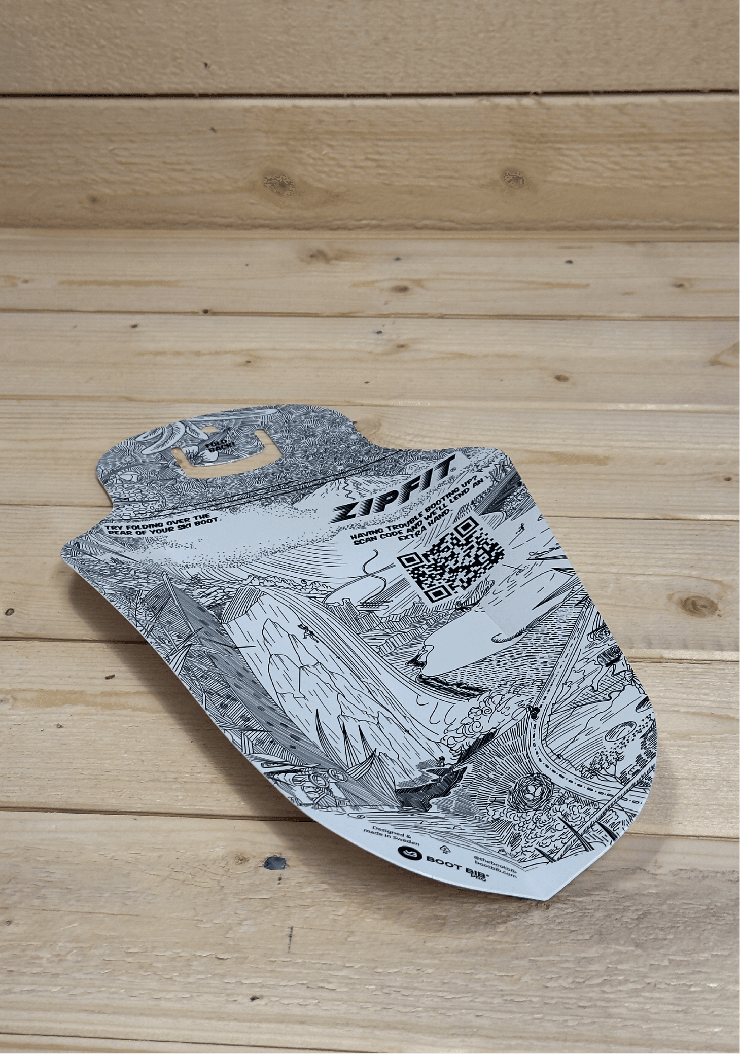 Bedruckte Verpackung in Form eines Wappenschildes, liegend auf hellem Holzgrund.  Die Grafik zeigt eine skizzierte Berglandschaft mit QR-Code.