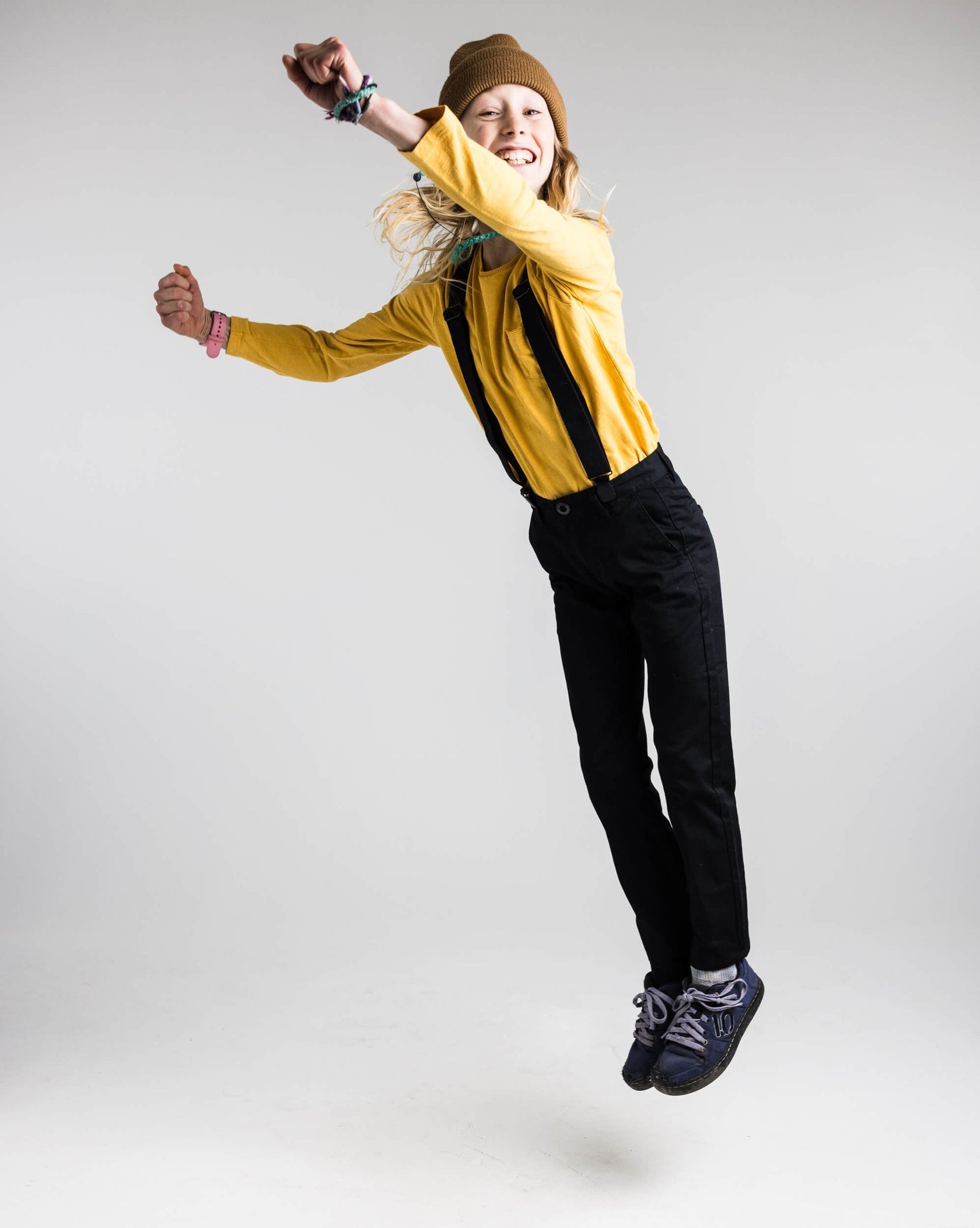 Ein junges Mädchen mit brauner Mütze springt vor einem weißen Hintergrund und reckt die Arme in die Luft.
