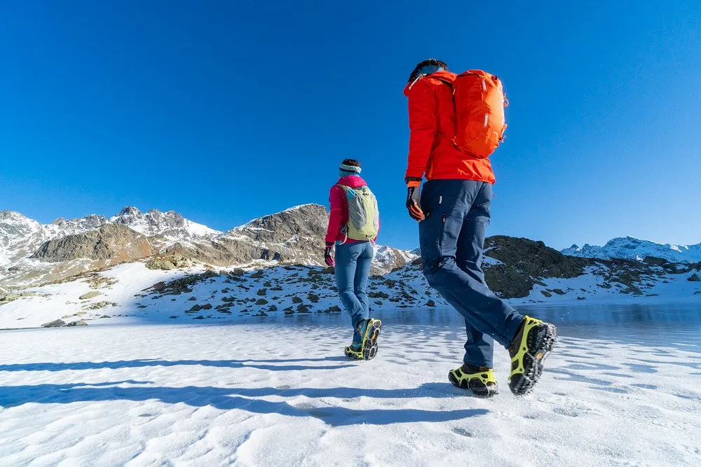 Zwei Personen mit Fast Schuhkrallen laufen auf Schnee vor Bergkulisse.