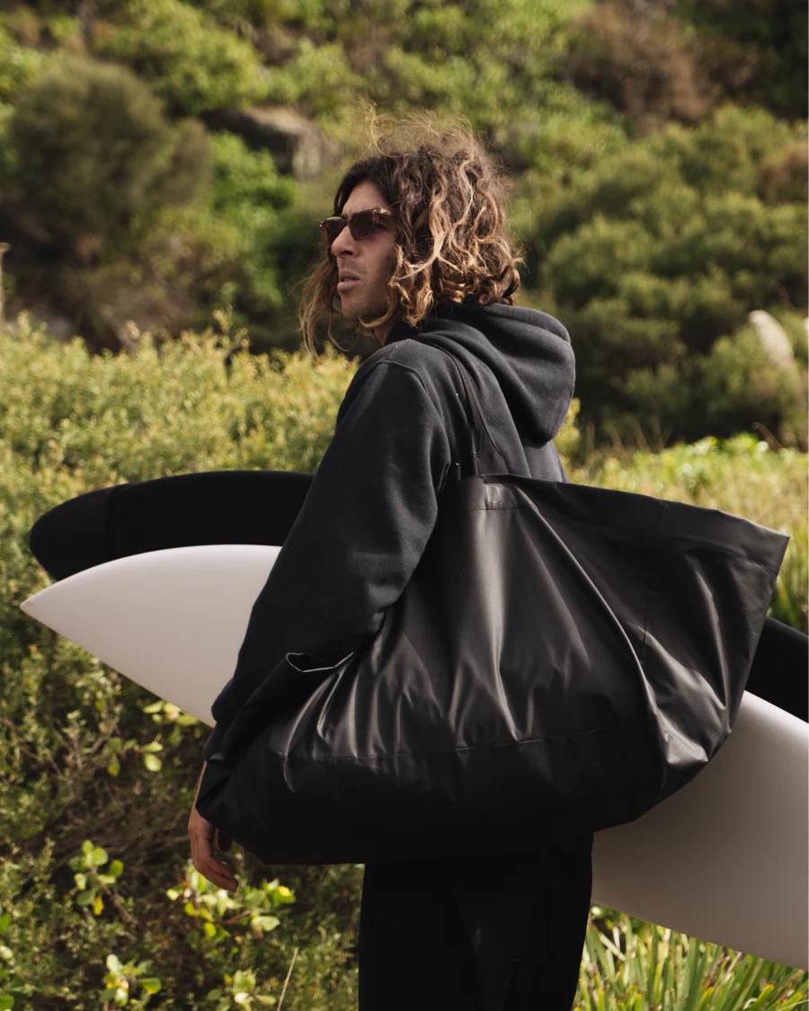 Surfer mit Tragetasche Mann mit Surfbrett und schwarzer Essential Tote Bag über der Schulter, im Hintergrund grüne Vegetation.