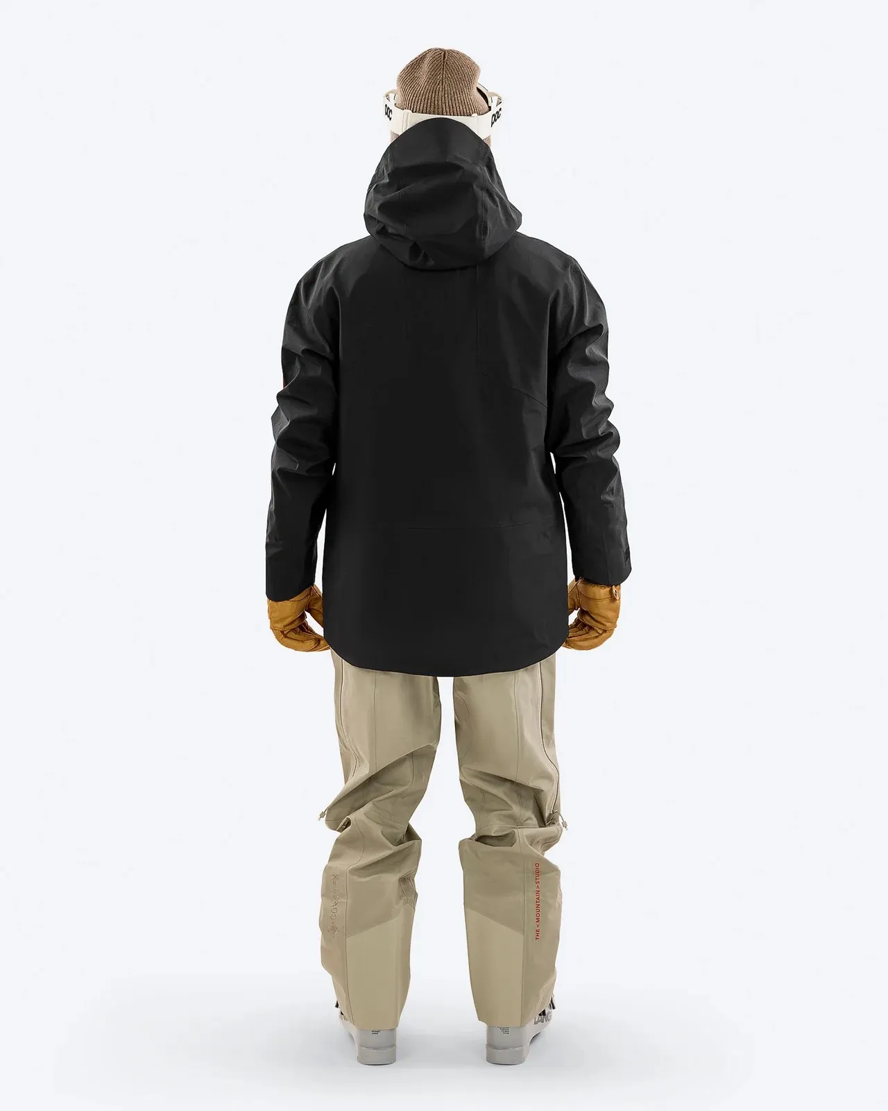 Rückansicht einer Person in Skibekleidung: Schwarze Jacke mit Kapuze, beigefarbene Hose, braune Handschuhe, Mütze und Skibrille.