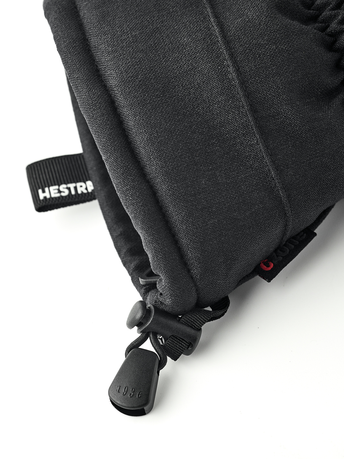 Schwarze Hestra-Fäustlinge Detailaufnahme eines schwarzen Hestra-Fäustlings mit Kordelzug und "1936" am Stopper. Ein Band mit der Aufschrift "Hestra" ist sichtbar.