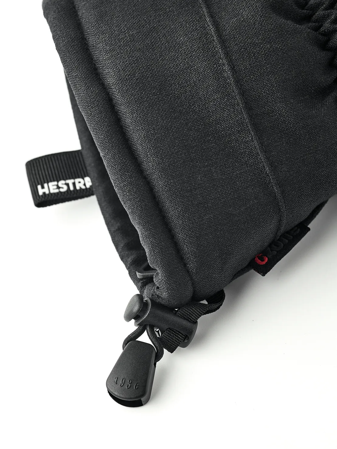 Detailaufnahme eines schwarzen Hestra-Fäustlings mit Kordelzug und "1936" am Stopper. Ein Band mit der Aufschrift "Hestra" ist sichtbar.