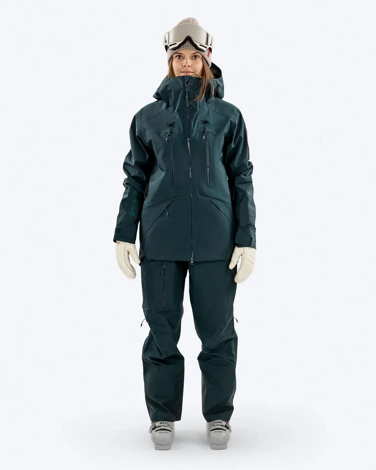 Eine Frau mit Skibrille und Mütze, gekleidet in einen dunkelgrünen Ski-Anzug mit Handschuhen, steht vor einem weißen Hintergrund.
