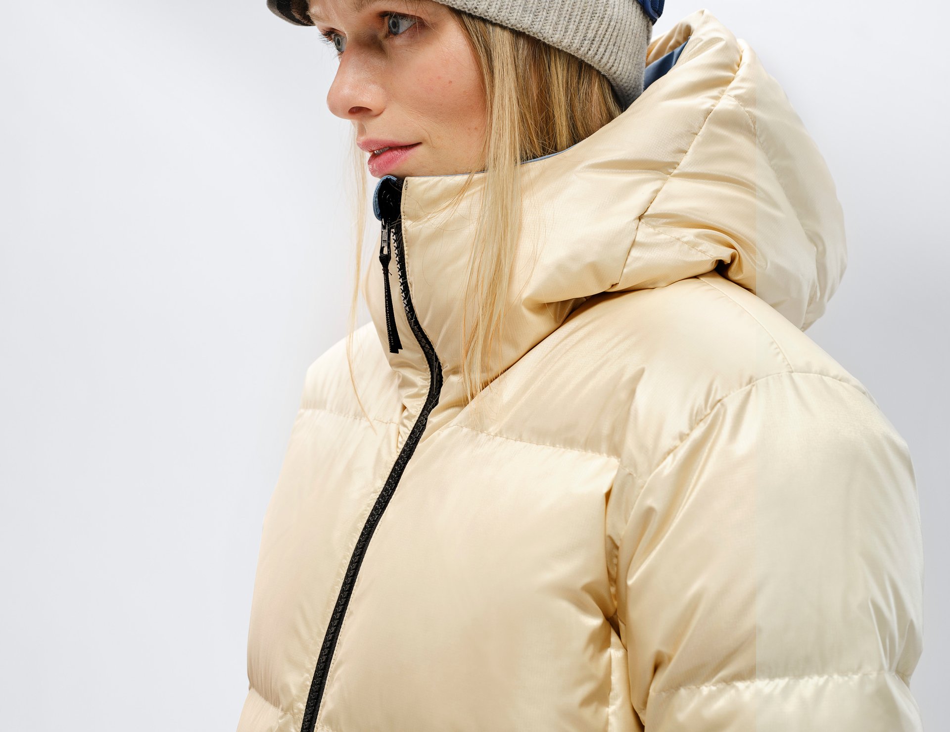Profil einer Frau mit beiger Daunenjacke und Mütze. Die Jacke hat einen schwarzen Reißverschluss und eine Kapuze.