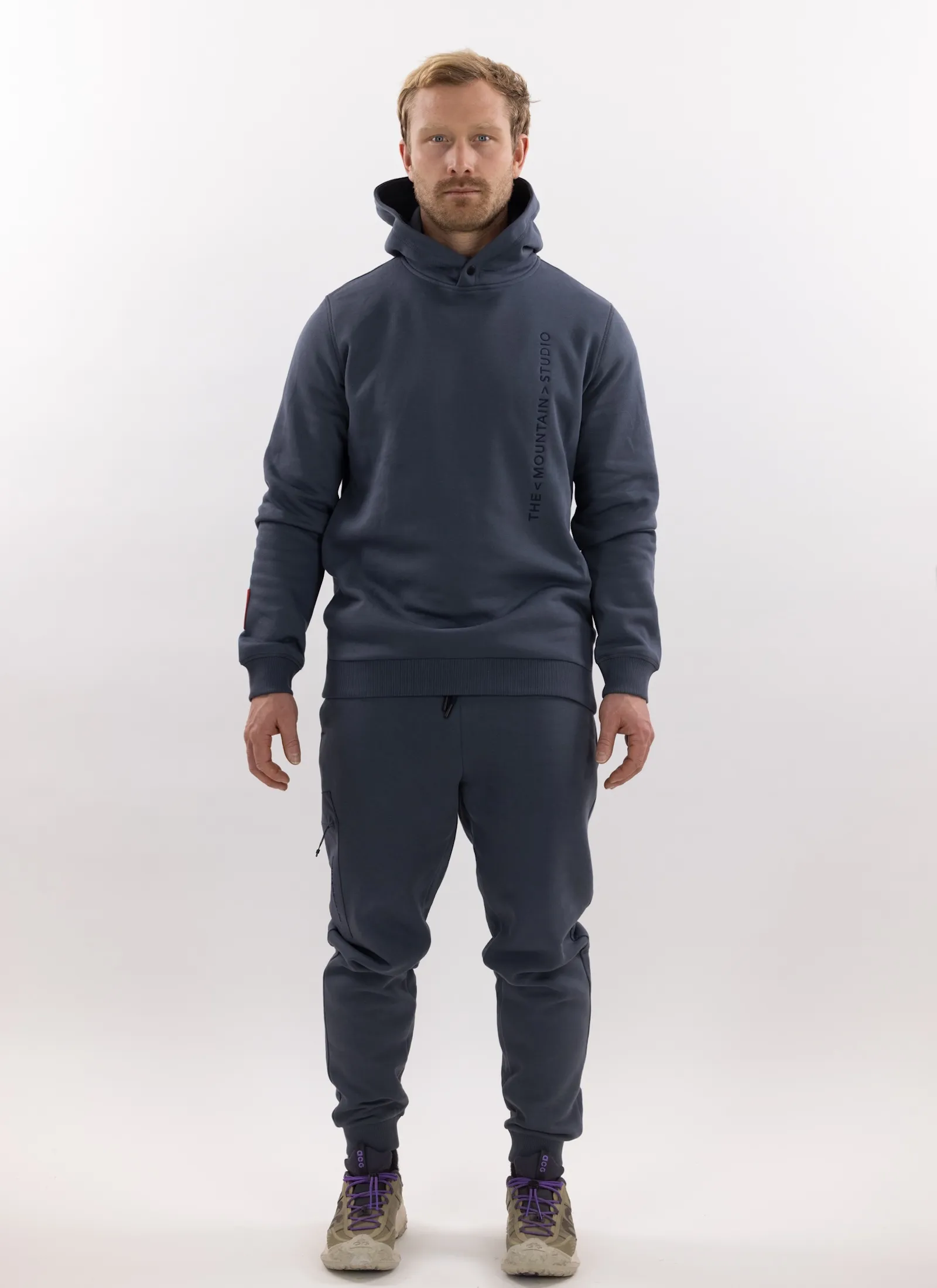 Mann in grauer Z-5 3L Hybrid Shell Jacket und passender Hose von The Mountain Studio vor weißem Hintergrund.