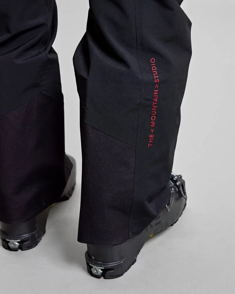 Detailansicht der schwarzen Y-1 HD GORE-TEX Pro 3L Shell Pant mit rotem "THE MOUNTAIN>STUDIO" Schriftzug und Skischuhen.