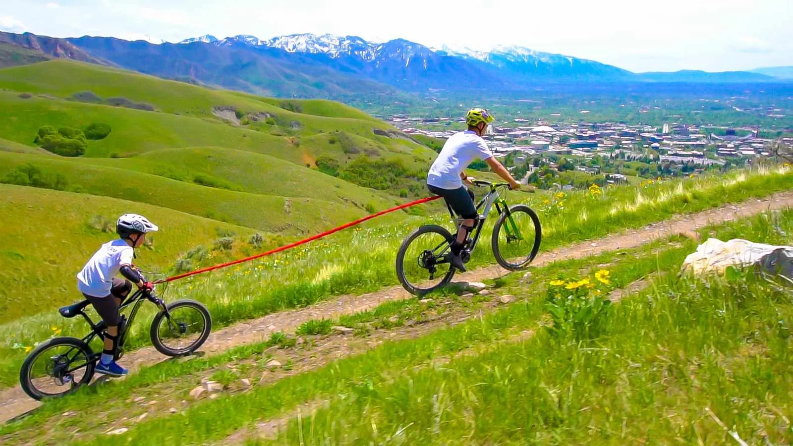 Zwei Kinder fahren mit dem Fahrrad bergauf, verbunden durch ein rotes Tow-Whee Abschleppseil vor einer grünen Hügellandschaft und Bergen.