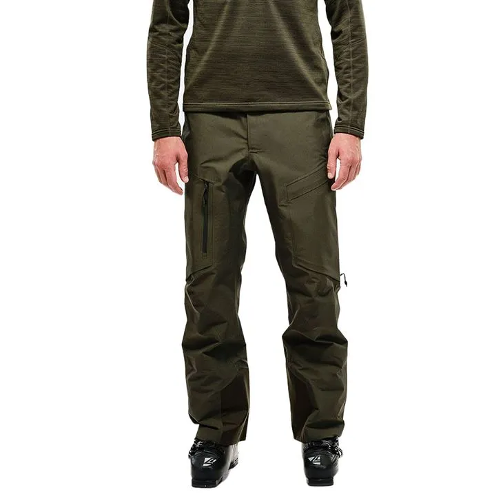 Mann in Skikleidung, trägt eine grüne Y-1 HD GORE-TEX Pro 3L Shell Pant und Skischuhe. Hose mit Taschen und Reißverschlüssen.