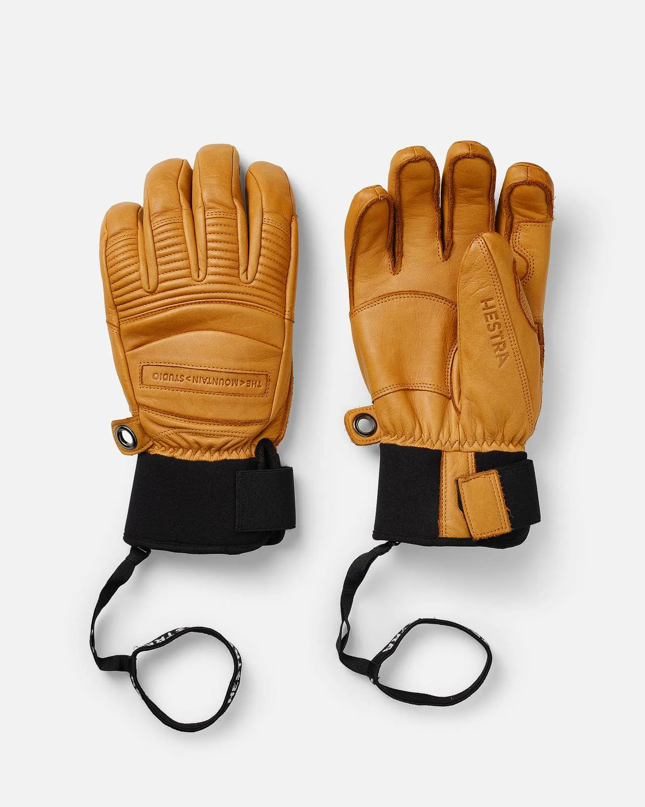 Zwei HESTRA® Lederhandschuhe in Sandfarbe mit schwarzen Neoprenmanschetten und Sicherheitsriemen auf weißem Hintergrund.