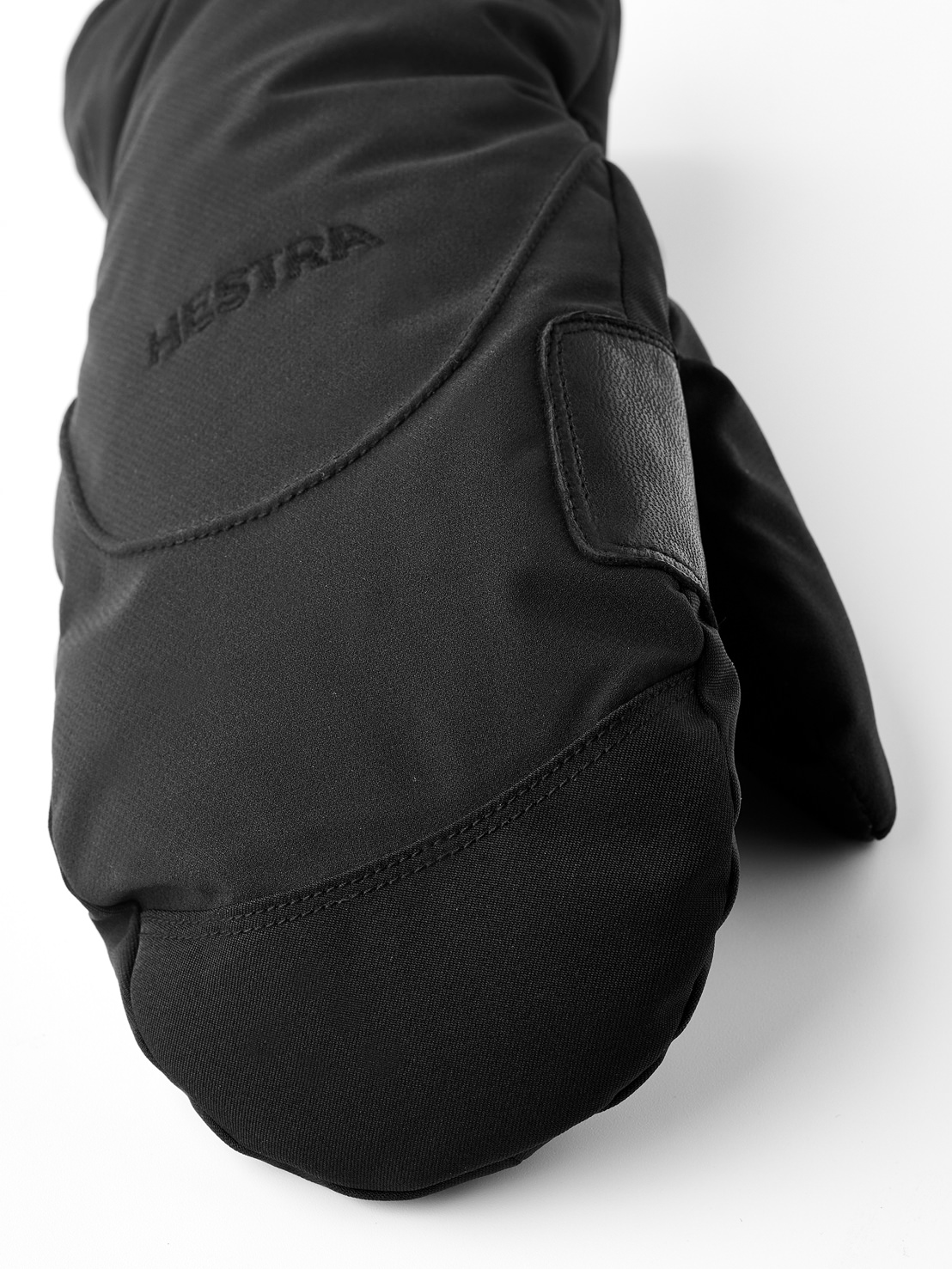 Schwarzer Fäustling mit Lederakzent Nahaufnahme eines schwarzen Fäustlings mit dezentem Hestra-Logo und Lederakzent auf hellem Hintergrund.