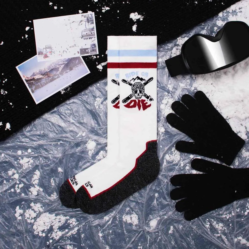 Weiße Kniestrümpfe mit "Ride or Die" Motiv neben Schneebrille, Handschuhen, Mütze und Winterfotos auf vereister Oberfläche.