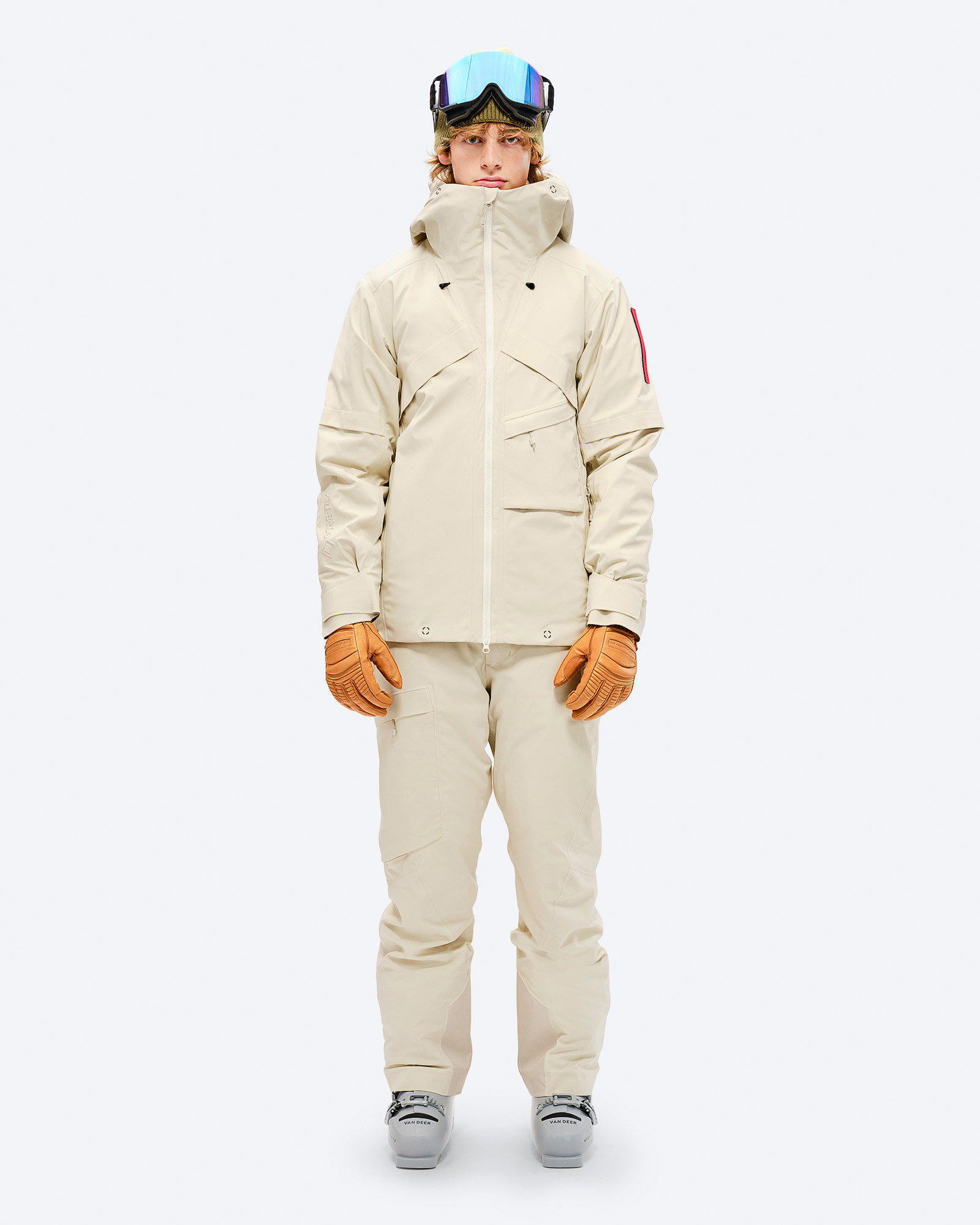 Person in beiger Ski-/Snowboardjacke mit GORE-TEX, Hose, Handschuhen und Skibrille vor weißem Hintergrund.