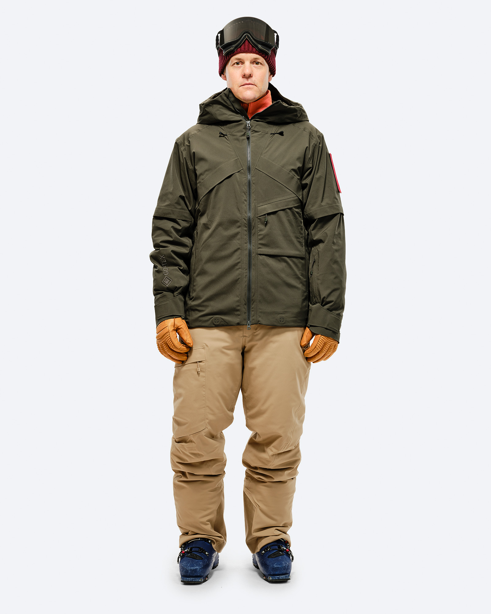 Person in Skikleidung: Olivgrüne Jacke, sandfarbene Hose und Skischuhe vor weißem Hintergrund.