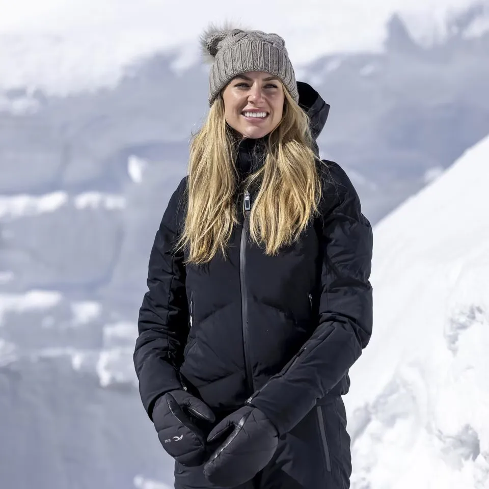 Eine Frau mit blonden, langen Haaren, trägt eine graue Mütze mit Bommel und eine schwarze Daunenjacke vor schneebedeckter Landschaft.