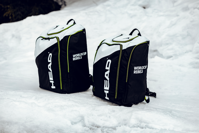 Zwei schwarze Head Rebels Racing Backpack L Rucksäcke liegen im Schnee. Sichtbar sind das Logo, der Schriftzug "Worldcup Rebels", Tragegriffe und gelbe Ziernähte.