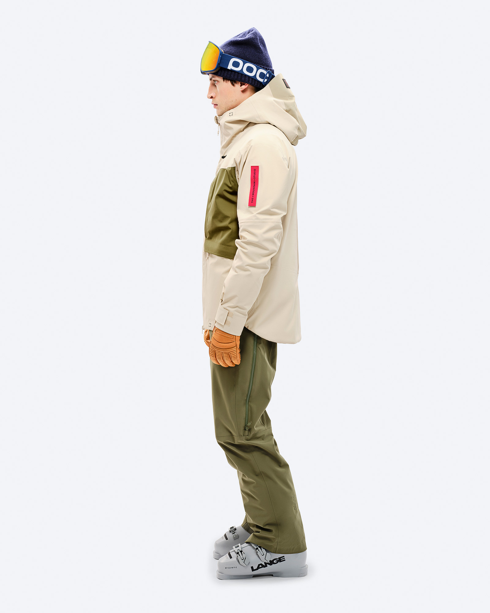 Mann in Ski-Kleidung, seitlich. Beige 3L Hybrid Shell Jacke, grüne Hose und Skischuhe. Blaue Mütze mit Skibrille.